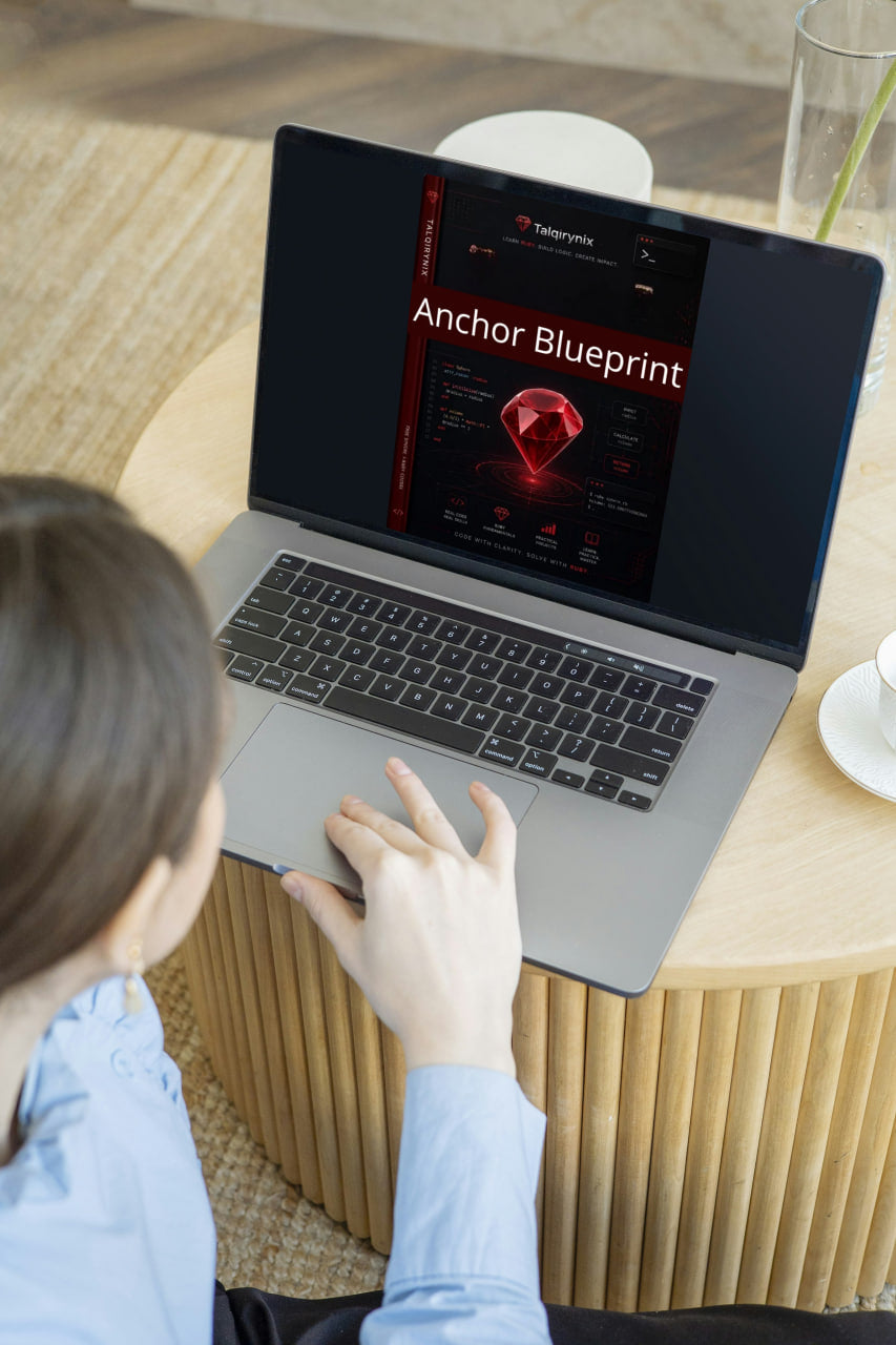 A person in a light blue shirt uses a silver laptop on a round wooden table. The laptop screen displays a dark interface with the text 'Anchor Blueprint' and a red gem graphic.